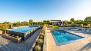 Vue d'un jardin moderne avec deux piscines, une semi-enterrée en acier et une enterrée, entourées de terrasses et verdure.