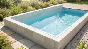 Piscine rectangulaire en blocs de polystyrène gris, remplie d'eau bleue claire, avec un jardin fleuri et une terrasse en bois.